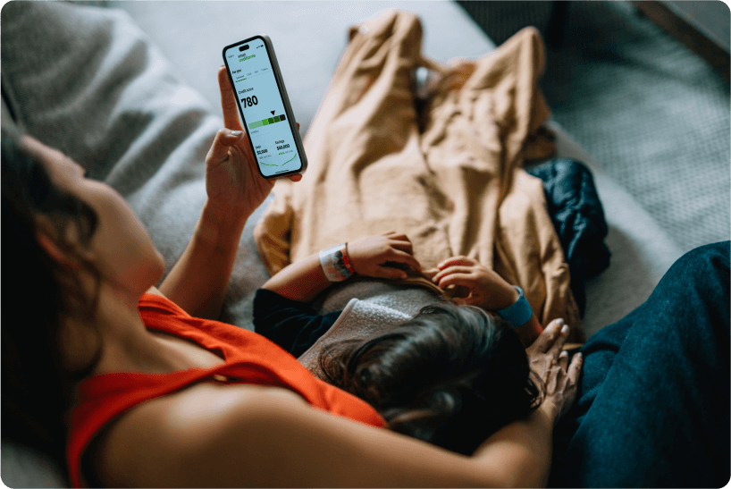 Woman on couch with child laying on her while she checks her cell phone with the Credit Karma app screen showing.