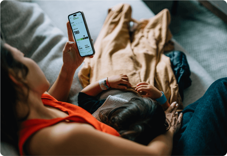 Woman on couch with child laying on her while she checks her cell phone with the Credit Karma app screen showing.